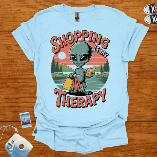 Light blue t-shirt with alien graphic and 'Shopping is my Therapy' text on a wooden surface.