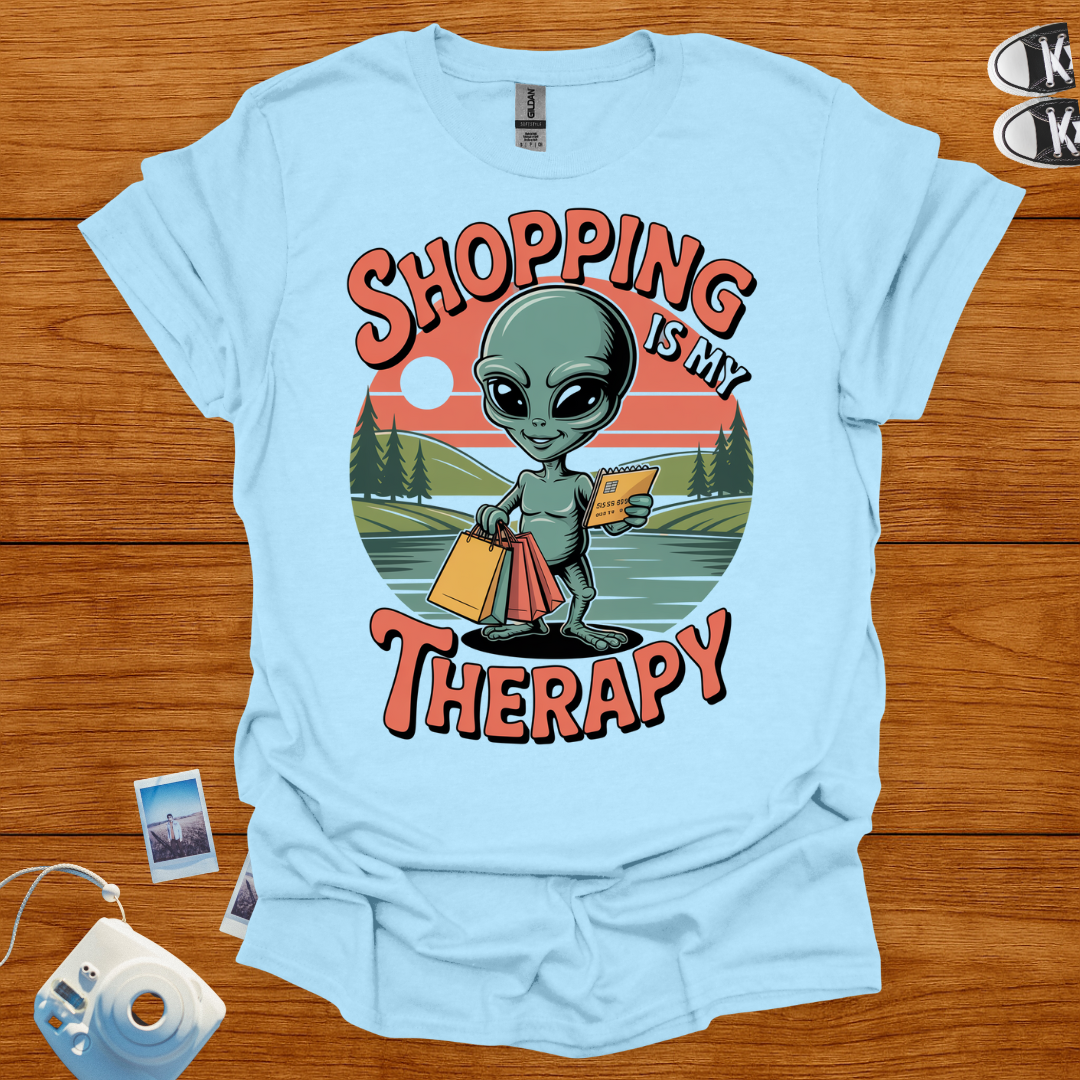 Light blue t-shirt with alien graphic and 'Shopping is my Therapy' text on a wooden surface.